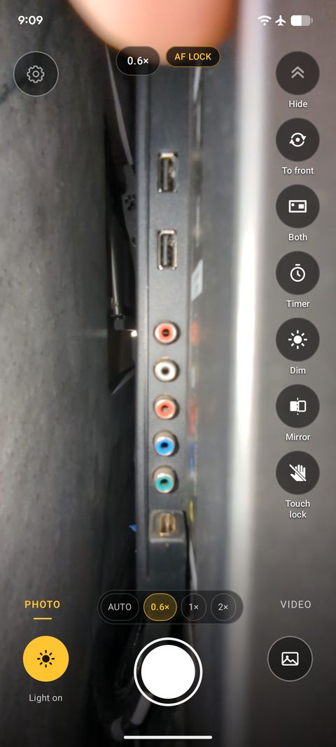 A real InspectCam screenshot behind a wall-mounted TV: the ports on its back lit by the phone's light, with a fingertip at the top of the frame.
